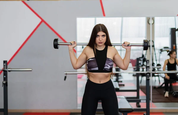 Hermosa chica se agacha con una barra en los hombros en el gimnasio . — Foto de Stock