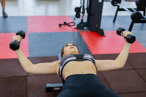 Beautiful girl trains pectoral muscles lying dumbbells on a bench in the gym.