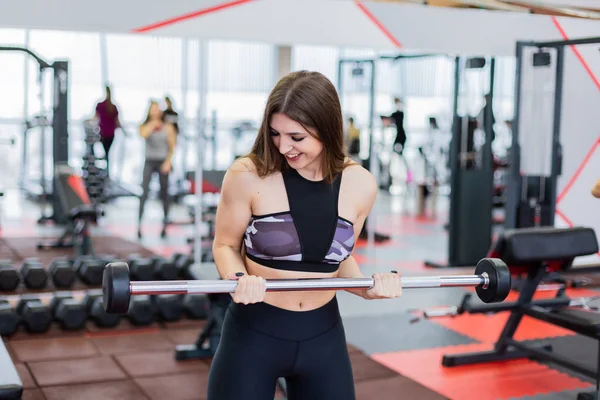 Hermosa chica entrena bíceps utilizando la barra en el suelo en el gimnasio . — Foto de Stock