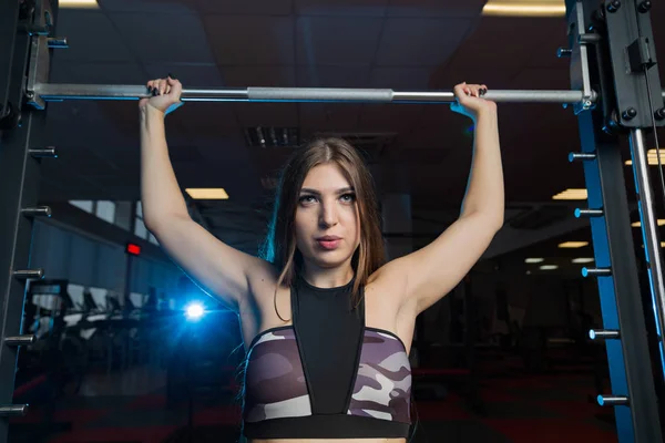 Hermosa sexy modelo de fitness sacude una barra de pesas sentado detrás de la cabeza en el gimnasio . — Foto de Stock