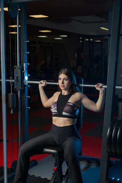 Hermosa sexy modelo de fitness sacude una barra de pesas sentado detrás de la cabeza en el gimnasio . — Foto de Stock