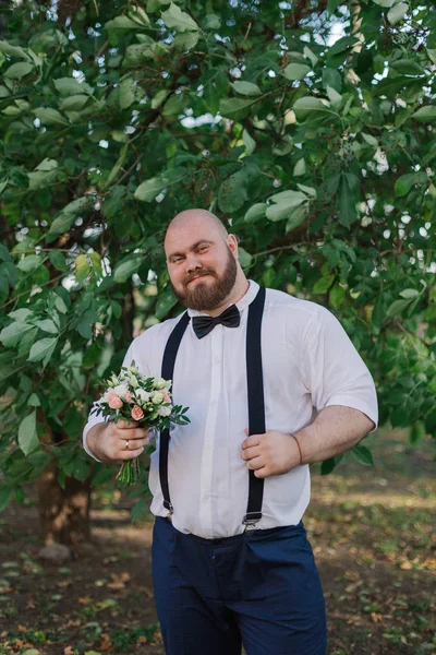 Stylish bearded fat groom with wedding bouquet in the park. — Stock Photo, Image