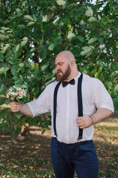 Stylish bearded fat groom with wedding bouquet in the park. — Stock Photo, Image