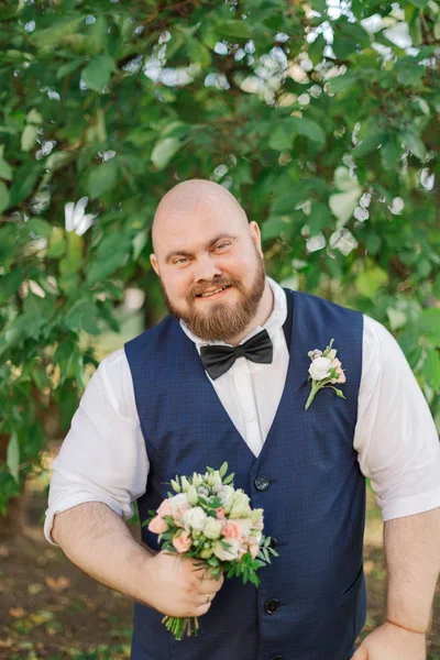 Stylish bearded fat groom with wedding bouquet in the park. — Stock Photo, Image