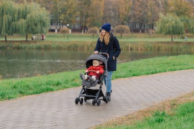 Bir kadın bir bebek arabası ve göl yakınındaki küçük bir çocuk ile parkta yürüyor.