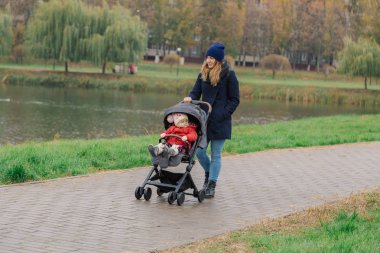 Bir kadın bir bebek arabası ve göl yakınındaki küçük bir çocuk ile parkta yürüyor.