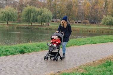 Bir kadın bir bebek arabası ve göl yakınındaki küçük bir çocuk ile parkta yürüyor.