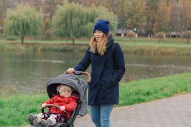 Bir kadın bir bebek arabası ve göl yakınındaki küçük bir çocuk ile parkta yürüyor.