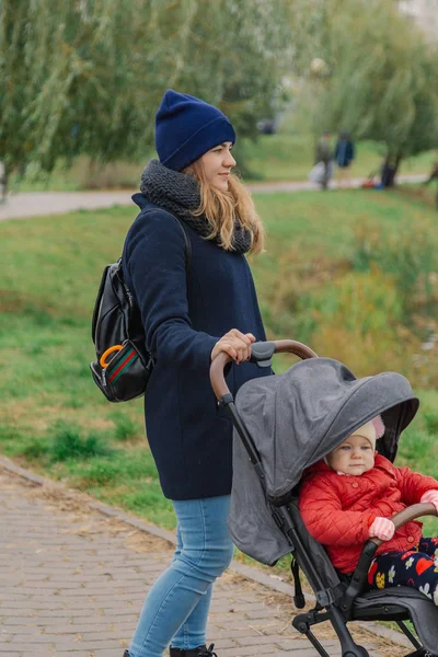 Una mujer camina en el parque con un cochecito y un niño pequeño cerca del lago . — Foto de Stock