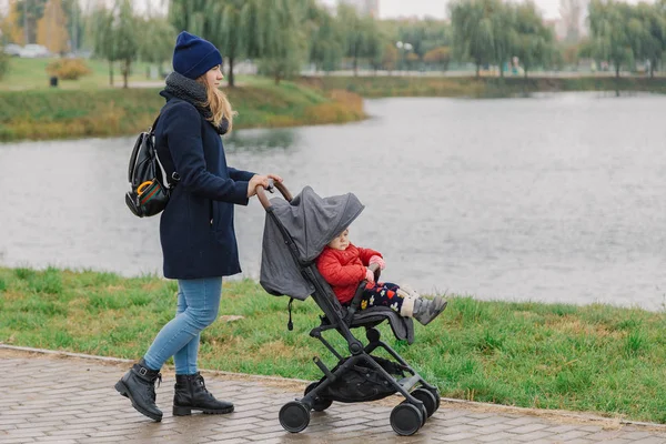 Una mujer camina en el parque con un cochecito y un niño pequeño cerca del lago . — Foto de Stock