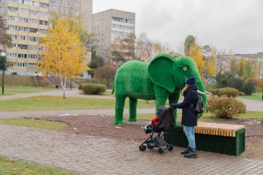 Bir kadın bir bebek arabası ve küçük bir çocuk ile parkta yürüyor.