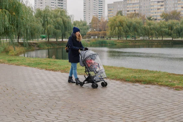 Una mujer camina en el parque con un cochecito y un niño pequeño cerca del lago . — Foto de Stock