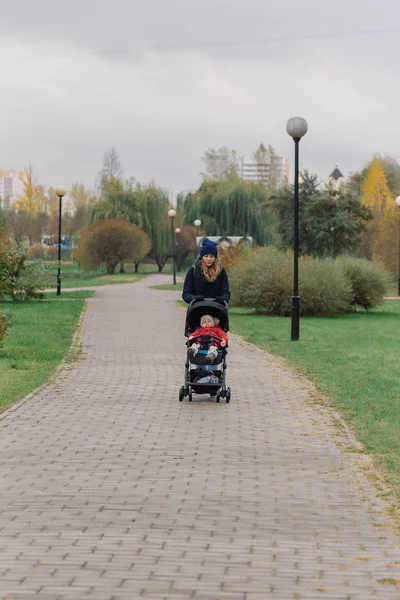 En kvinna går i parken med en barnvagn och ett litet barn. — Stockfoto