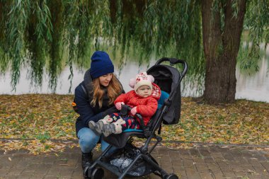 Bir kadın bir bebek arabası ve göl yakınındaki küçük bir çocuk ile parkta yürüyor.