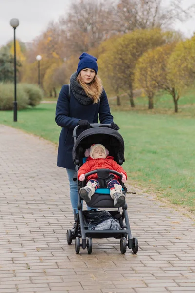 Una mujer camina por el parque con un cochecito y un niño . — Foto de Stock