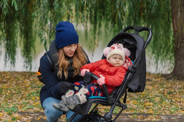 Una mujer camina en el parque con un cochecito y un niño pequeño cerca del lago . — Foto de Stock