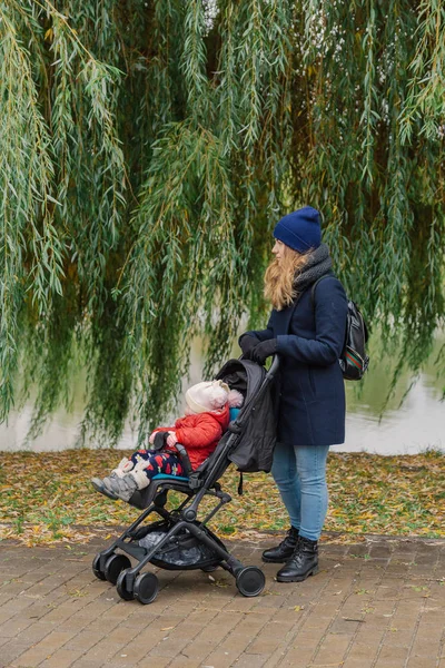 Una mujer camina en el parque con un cochecito y un niño pequeño cerca del lago . — Foto de Stock