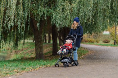 Bir kadın bir bebek arabası ve küçük bir çocuk ile parkta yürüyor.