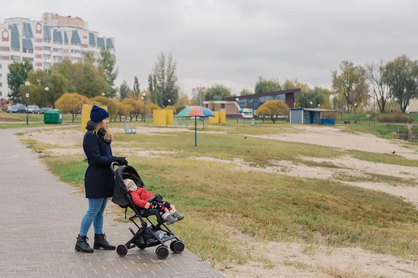 Uma mulher caminha no parque com um carrinho e uma criança pequena perto do lago . — Fotografia de Stock