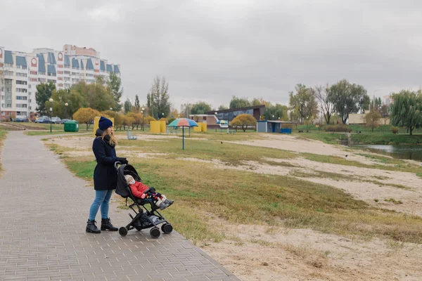 Uma mulher caminha no parque com um carrinho e uma criança pequena perto do lago . — Fotografia de Stock