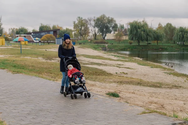 Una mujer camina en el parque con un cochecito y un niño pequeño cerca del lago . — Foto de Stock