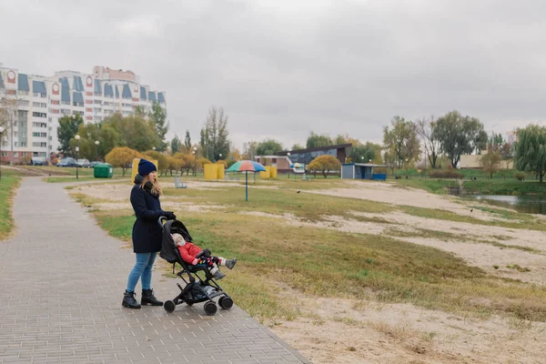 Uma mulher caminha no parque com um carrinho e uma criança pequena perto do lago . — Fotografia de Stock