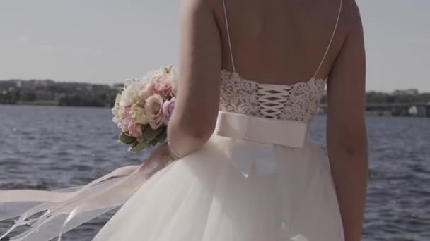 À mi-chemin d'une charmante mariée tenant un délicat bouquet de mariage. Debout près du lac et regardant sur la distance par une journée nuageuse . — Video