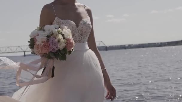 À mi-chemin d'une charmante mariée tenant un délicat bouquet de mariage. Debout près du lac et regardant sur la distance par une journée nuageuse . — Video