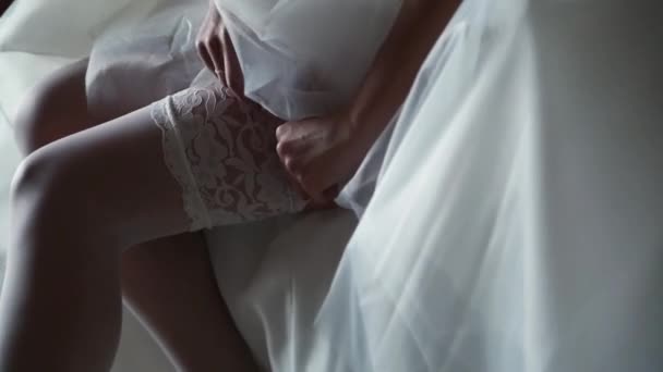 Η νύφη φοράει μια λευκή καλτσοδέτα στο πόδι της, κάθεται στο κρεβάτι. — Αρχείο Βίντεο