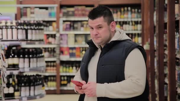 Gordo no supermercado está no departamento com álcool e olha para o telefone . — Vídeo de Stock