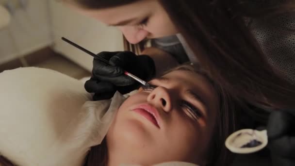 Ein sehr schönes Mädchen in einem Schönheitssalon macht eine Laminierung Wimpern. Kosmetikerin streicht Farbe auf die Wimpern. — Stockvideo