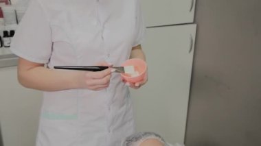 Bir güzellik uzmanı yüz maskesini fırçayla karıştırır. Genç kozmetikçi kozmetik kremini küçük bir kapta karıştırıyor. Kozmetik kremi karıştırılıyor.