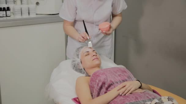 Geschoolde schoonheidsspecialist zet masker op gezicht van liggende vrouw in spa salon binnenshuis. — Stockvideo