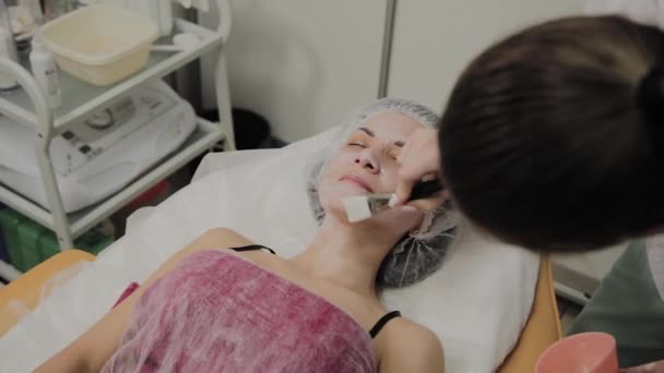 Cosmétologue qualifié met masque sur le visage de la femme couchée dans le salon de spa à l'intérieur . — Video