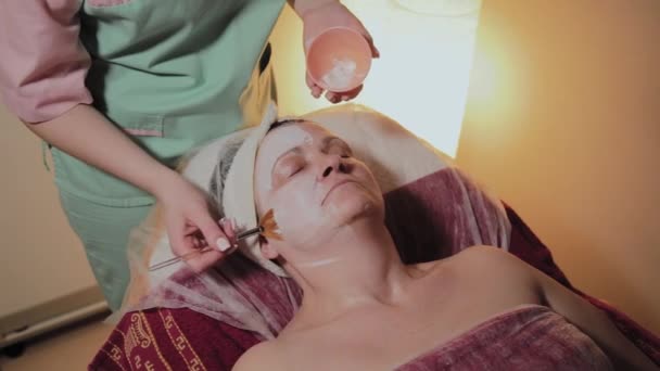 Een professionele schoonheidsspecialist past een gezichtsmasker aan een bejaarde vrouw toe. Cosmetische innovaties. — Stockvideo