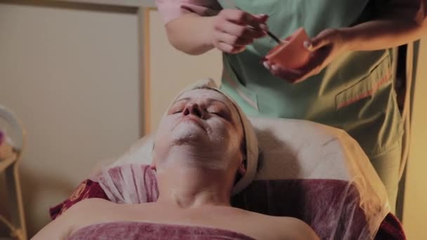 Profesjonalny kosmetolog stosuje maskę twarzy do starszej kobiety. Innowacje kosmetyczno-kosmetyczne. — Wideo stockowe
