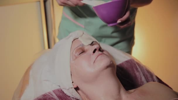 Um cosmetologista profissional aplica uma máscara facial a uma mulher idosa. Inovações cosmológicas . — Vídeo de Stock