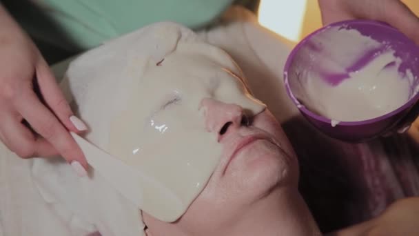 Ένας επαγγελματίας αισθητικός εφαρμόζει μια μάσκα προσώπου σε μια ηλικιωμένη γυναίκα. Καλλυννολογικές καινοτομίες. — Αρχείο Βίντεο
