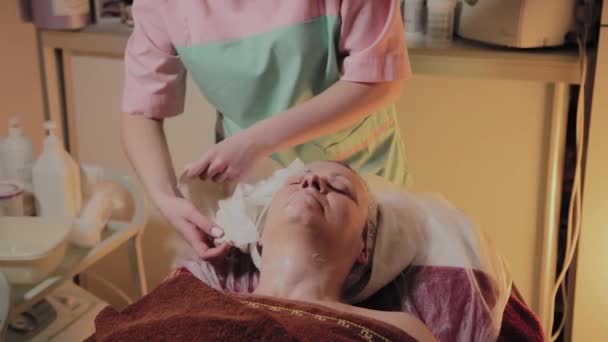 Professionele schoonheidsspecialist verwijdert het masker van het gezicht van een oudere vrouw. Cosmetische innovaties. — Stockvideo