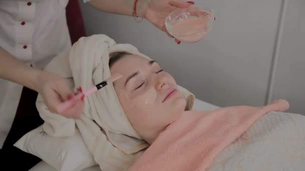 Um cosmetologista profissional aplica uma máscara facial a uma jovem com um pincel. Novo conceito em cosmetologia . — Vídeo de Stock