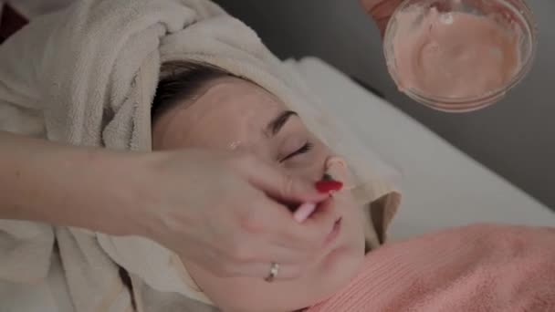 Um cosmetologista profissional aplica uma máscara facial a uma jovem com um pincel. Novo conceito em cosmetologia . — Vídeo de Stock