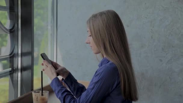 Menina bonita sentada à janela com um telefone e uma bebida na mesa . — Vídeo de Stock
