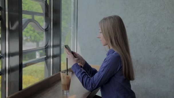 Hermosa chica sentada junto a la ventana con un teléfono y una bebida en la mesa . — Vídeos de Stock