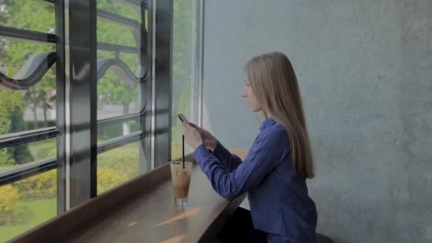 Hermosa chica sentada junto a la ventana con un teléfono y una bebida en la mesa . — Vídeos de Stock
