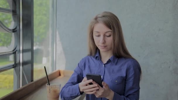 Hermosa chica sentada junto a la ventana con un teléfono en un café . — Vídeo de stock