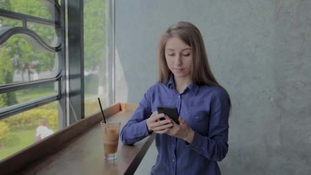 Schönes Mädchen sitzt am Fenster mit einem Telefon in einem Café. — Stockvideo