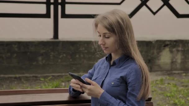 Hermosa chica feliz sentado en un banco en un parque de la ciudad con un teléfono móvil . — Vídeos de Stock