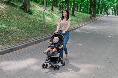 Bebek arabasında çocuklu güzel kadın bir yaz parkında yürüyor.