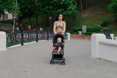 Bebek arabasında çocuklu güzel kadın bir yaz parkında yürüyor.