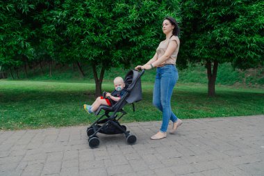 Bebek arabasında çocuklu güzel kadın bir yaz parkında yürüyor.